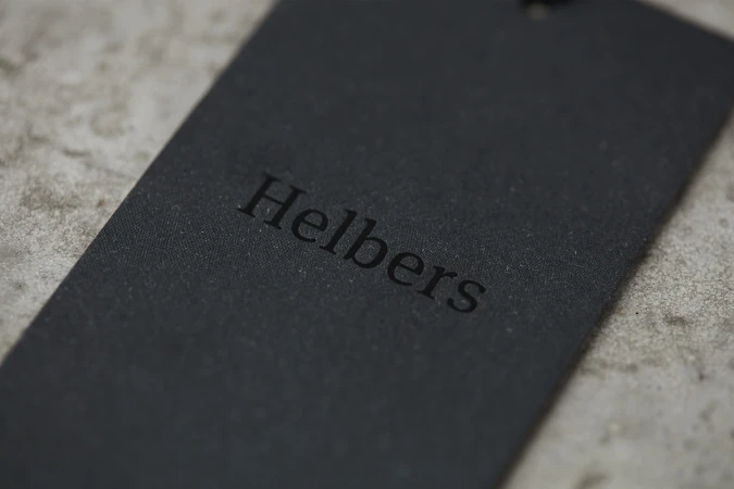 Black paper clothing tag with embossed Helbers brand logo resting on a textured concrete surface.