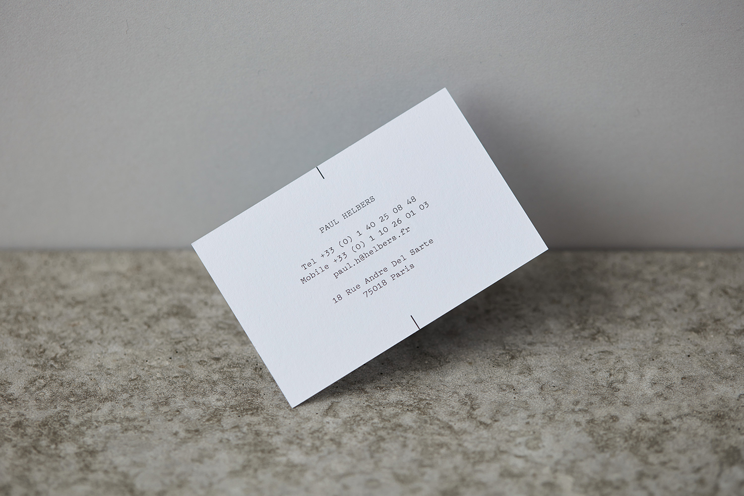 White business card with typewriter text resting at an angle on a textured stone and plain wall background.