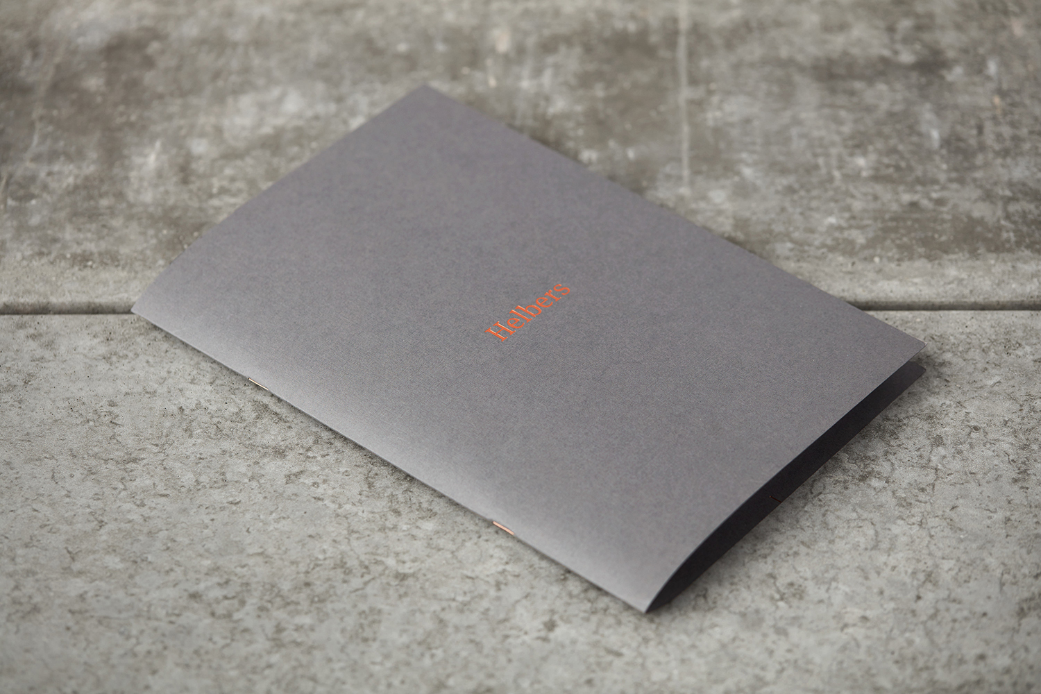 A dark grey folded paper booklet with embossed orange Helbers branding resting on a textured stone surface