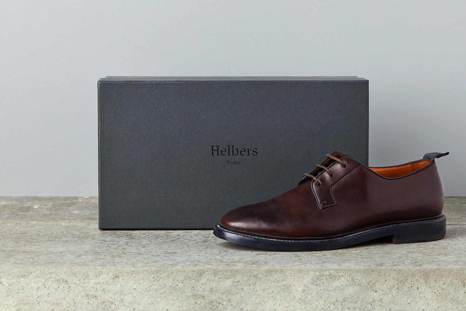 Dark brown leather derby shoe resting on a textured surface beside a dark grey Helbers Paris shoebox against a neutral background.