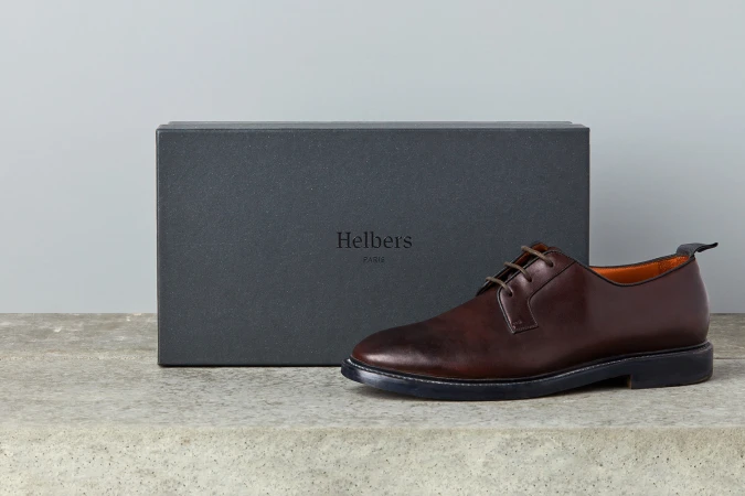 Dark brown leather derby shoe resting on a textured surface beside a dark grey Helbers Paris shoebox against a neutral background.