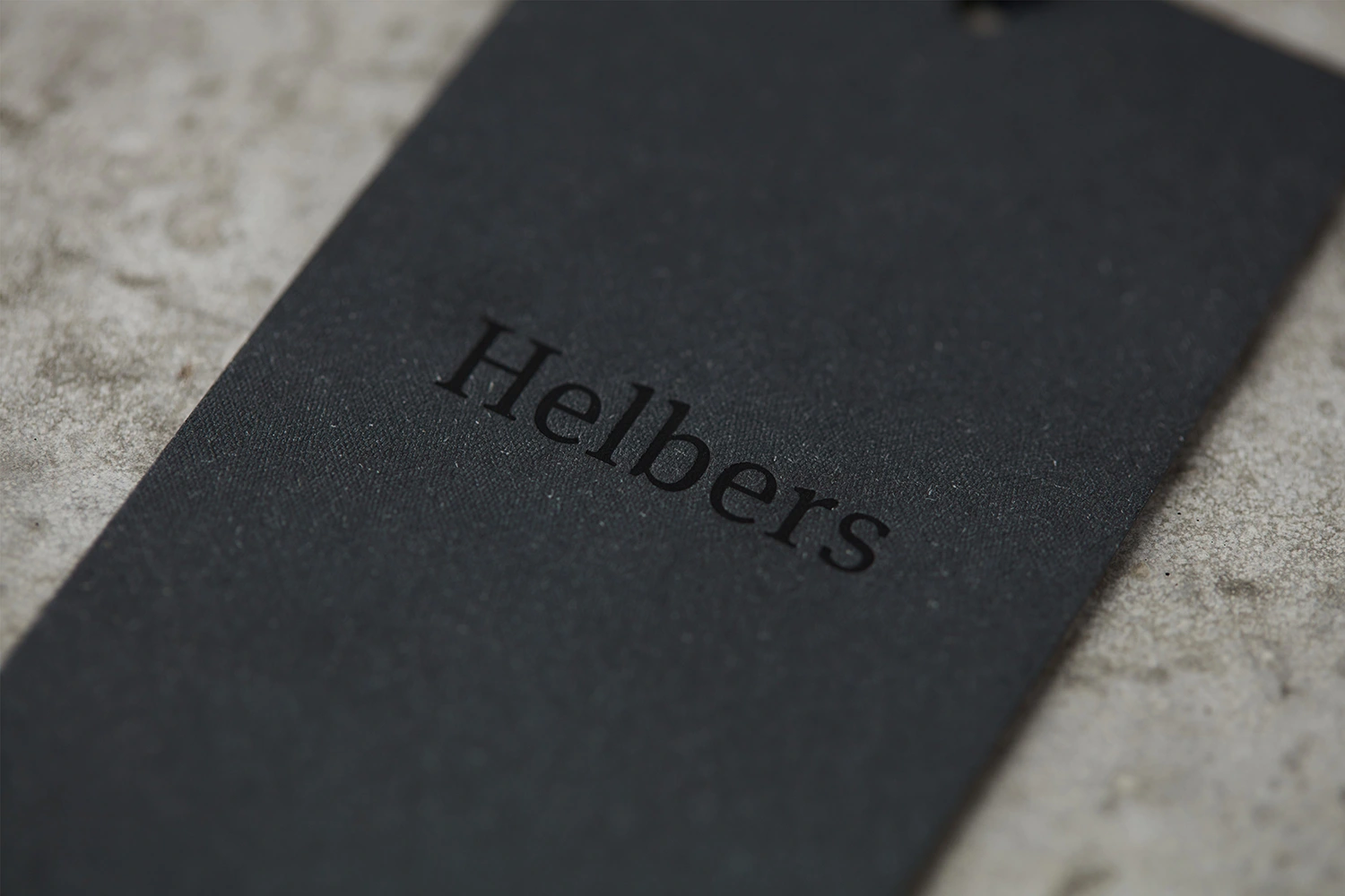 Black paper clothing tag with embossed Helbers brand logo resting on a textured concrete surface.