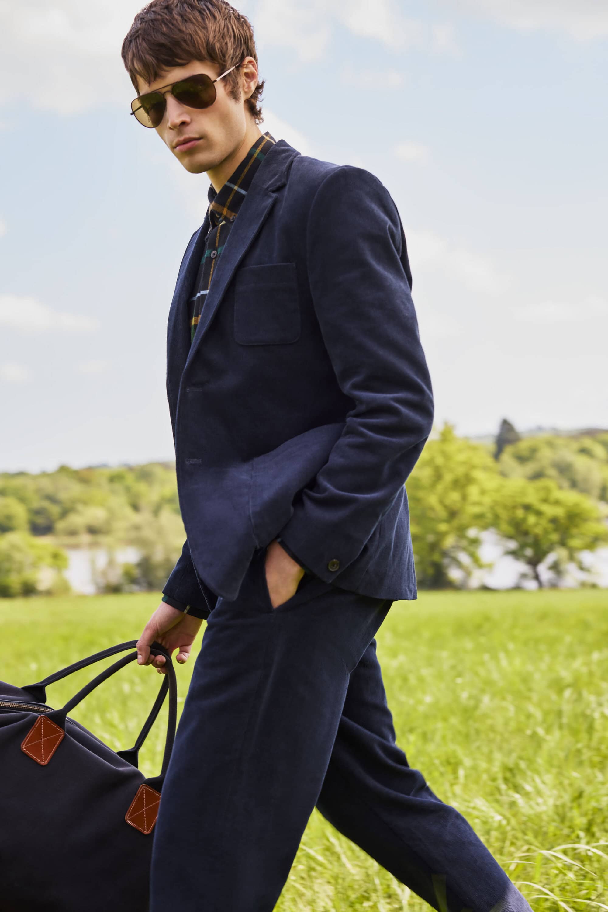 A young male model walking through a grassy field wearing a dark navy blazer, trousers, patterned shirt, and sunglasses, carrying a duffel bag.