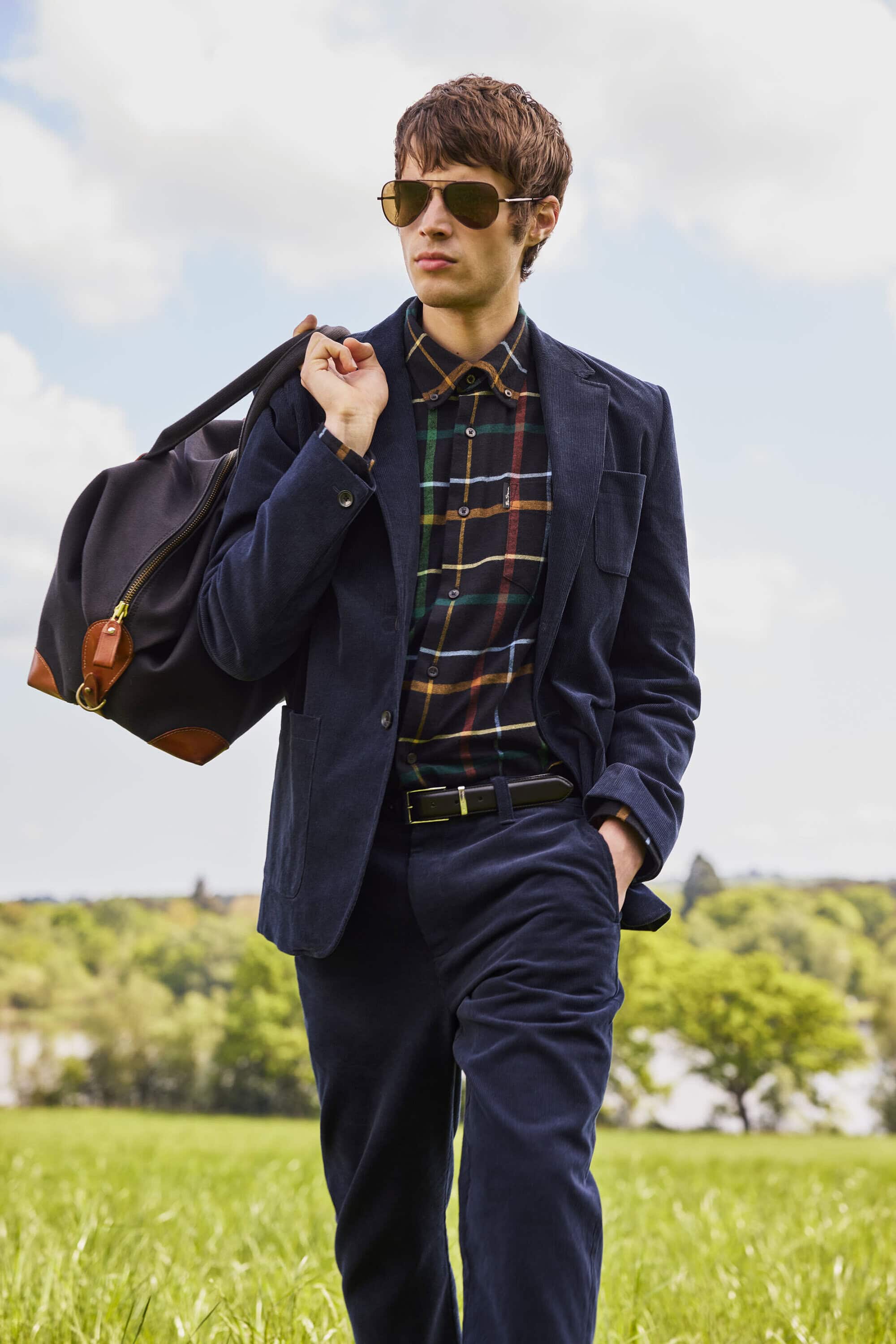 Young man in dark blue suit and plaid shirt carrying a duffel bag across a green meadow under an overcast sky.