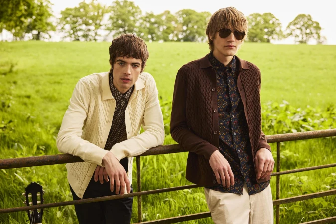 Two male models leaning on a wooden fence in a green meadow wearing patterned shirts and cardigans for Ben Sherman Fall 2025 campaign.