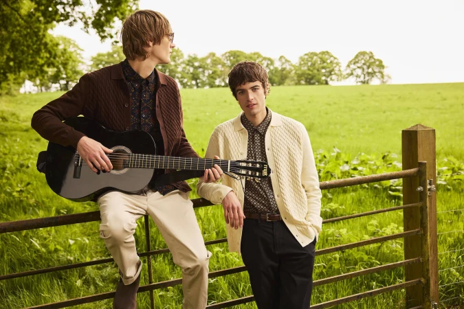 Two men posing outdoors leaning on a wooden fence in a green meadow holding a black acoustic guitar