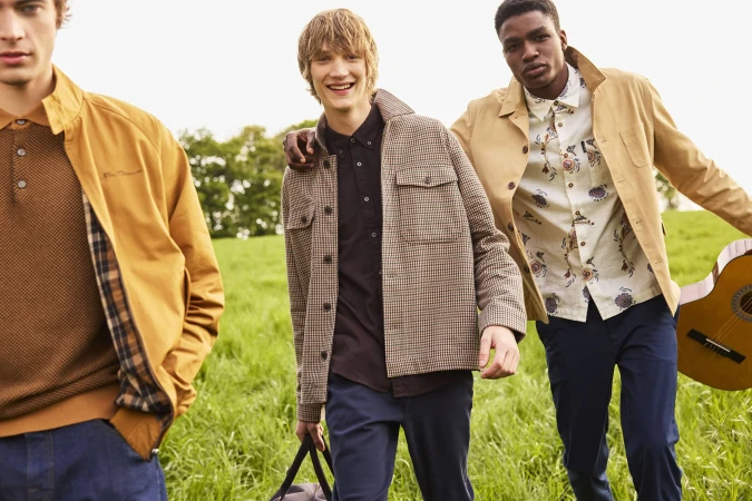 Three young men walking through a green meadow, wearing casual autumn jackets and carrying a guitar and bag for Ben Sherman.