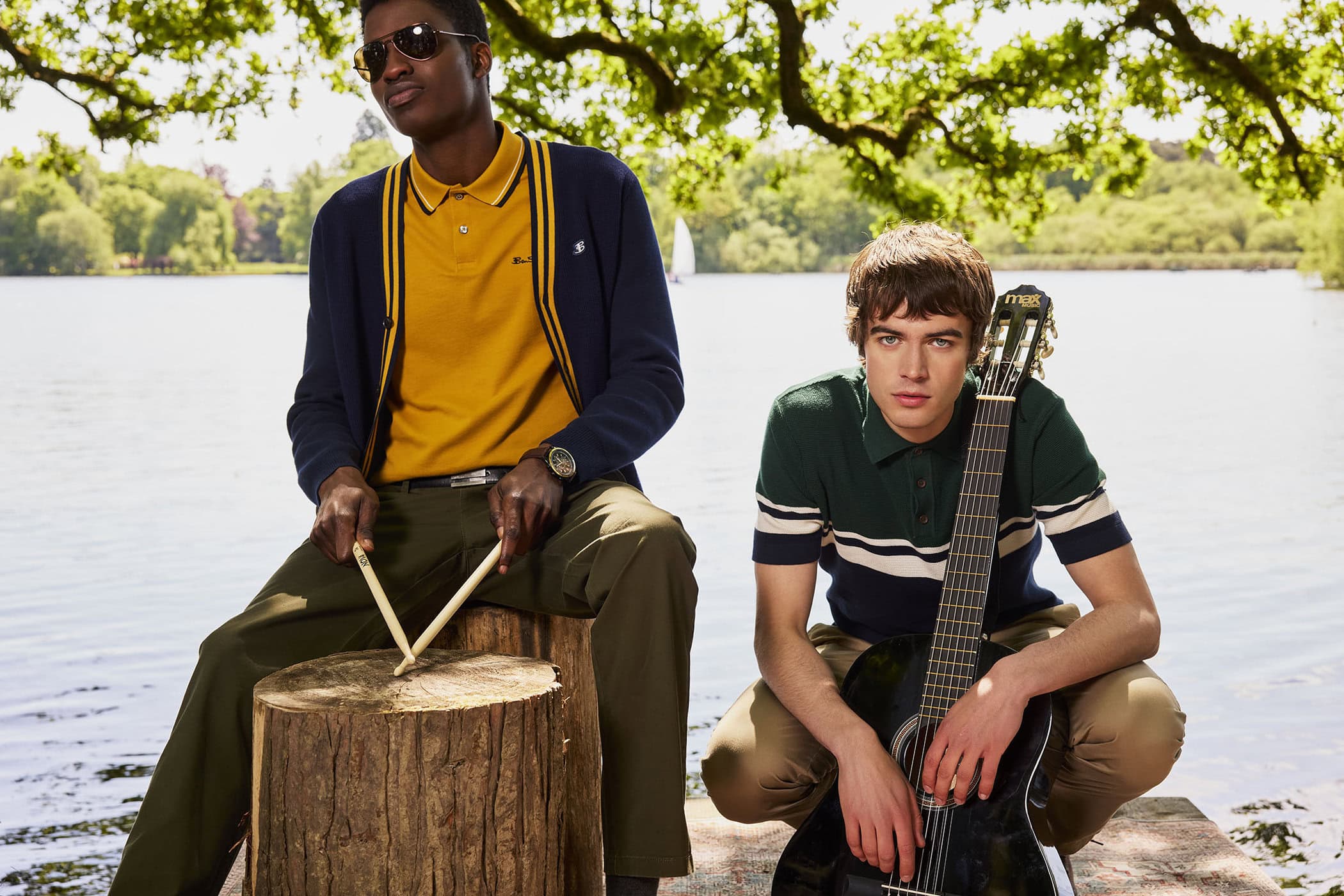 Two male models posing outdoors by a lake, one holding drumsticks on a tree stump and the other holding an acoustic guitar.