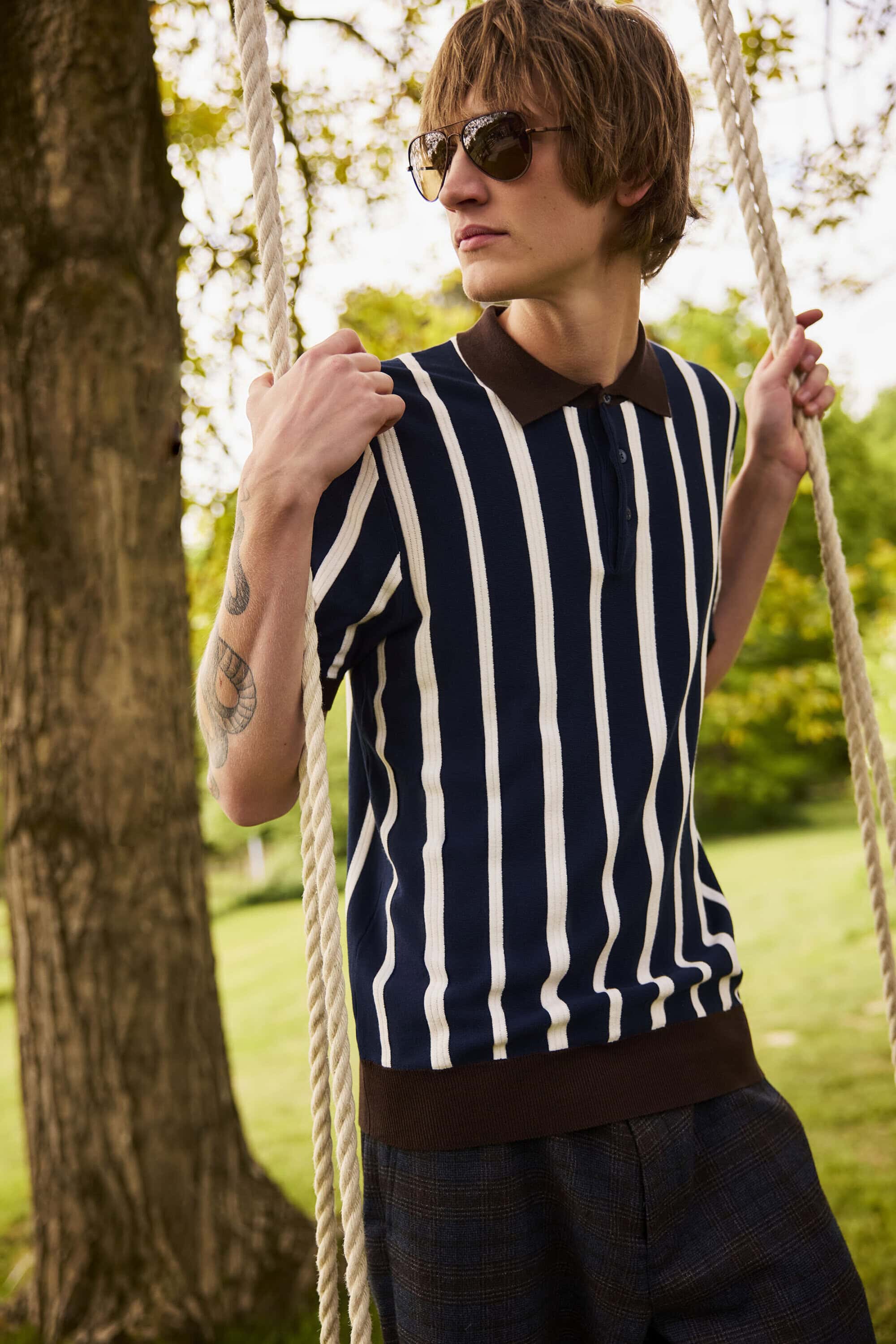 Young man in a striped polo shirt wearing sunglasses while holding rope on an outdoor swing in a park