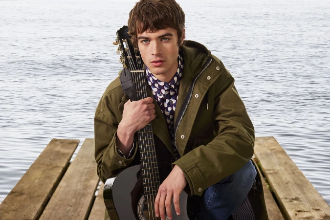Young man in olive green hooded jacket holding an acoustic guitar on a wooden dock over water.