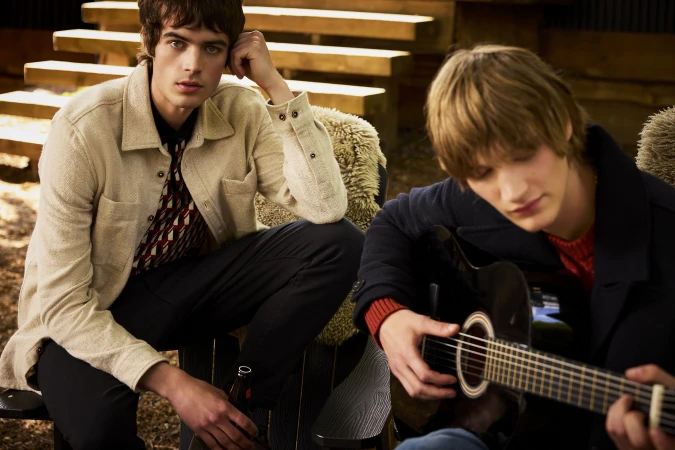 Two young male models seated outdoors, one holding a guitar and the other holding a bottle