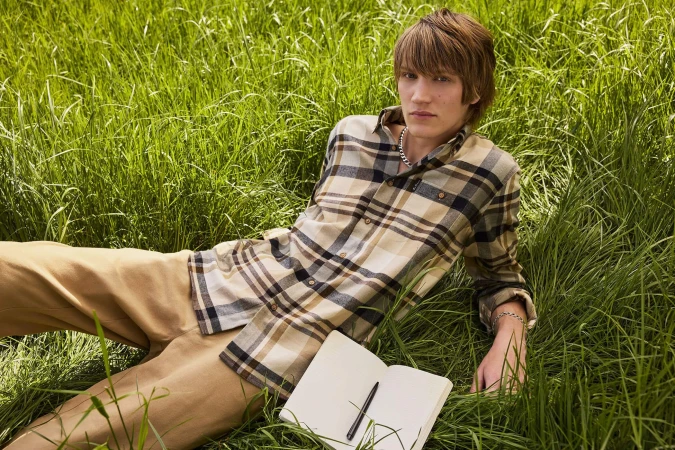 A young male model lying in tall green grass wearing a plaid shirt and tan trousers, with an open notebook nearby.