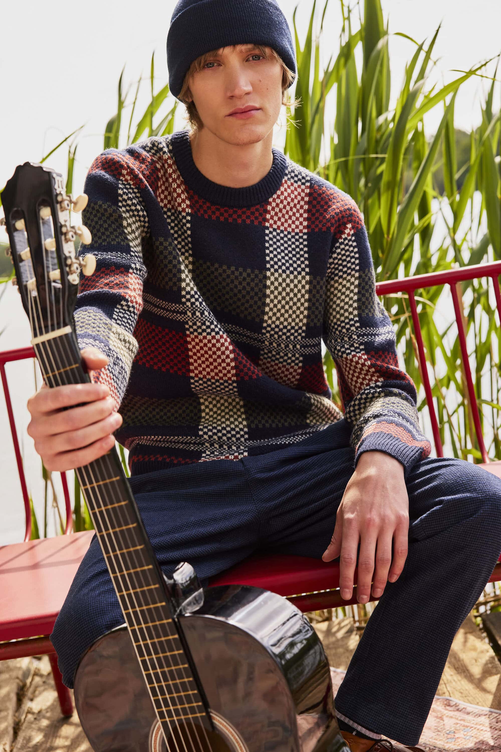 Young male model holding acoustic guitar wearing patterned sweater and beanie outdoors by tall green reeds