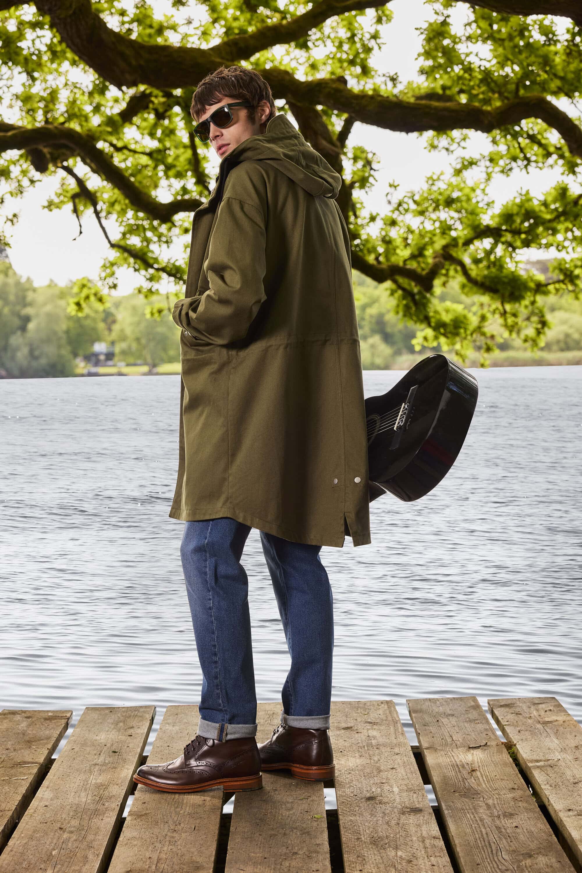 Man wearing a long olive green parka, blue jeans, and brown leather shoes standing on a wooden pier holding a black guitar