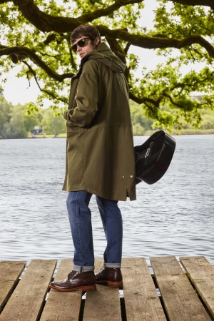 Man wearing a long olive green parka, blue jeans, and brown leather shoes standing on a wooden pier holding a black guitar