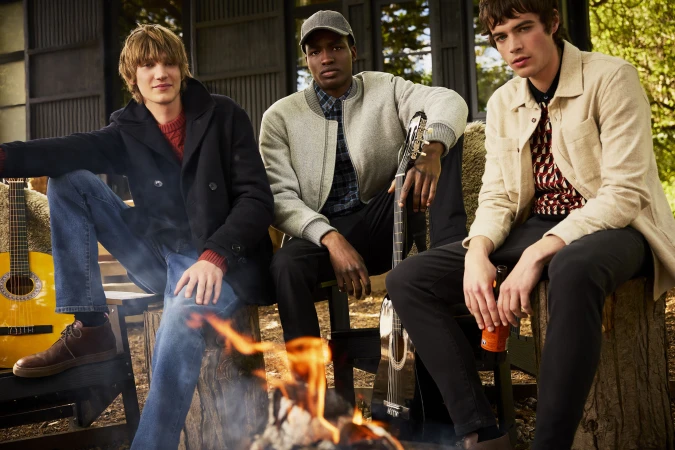 Three male models seated outdoors around a campfire holding acoustic guitars for the Ben Sherman Signature fall 2025 ad campaign.