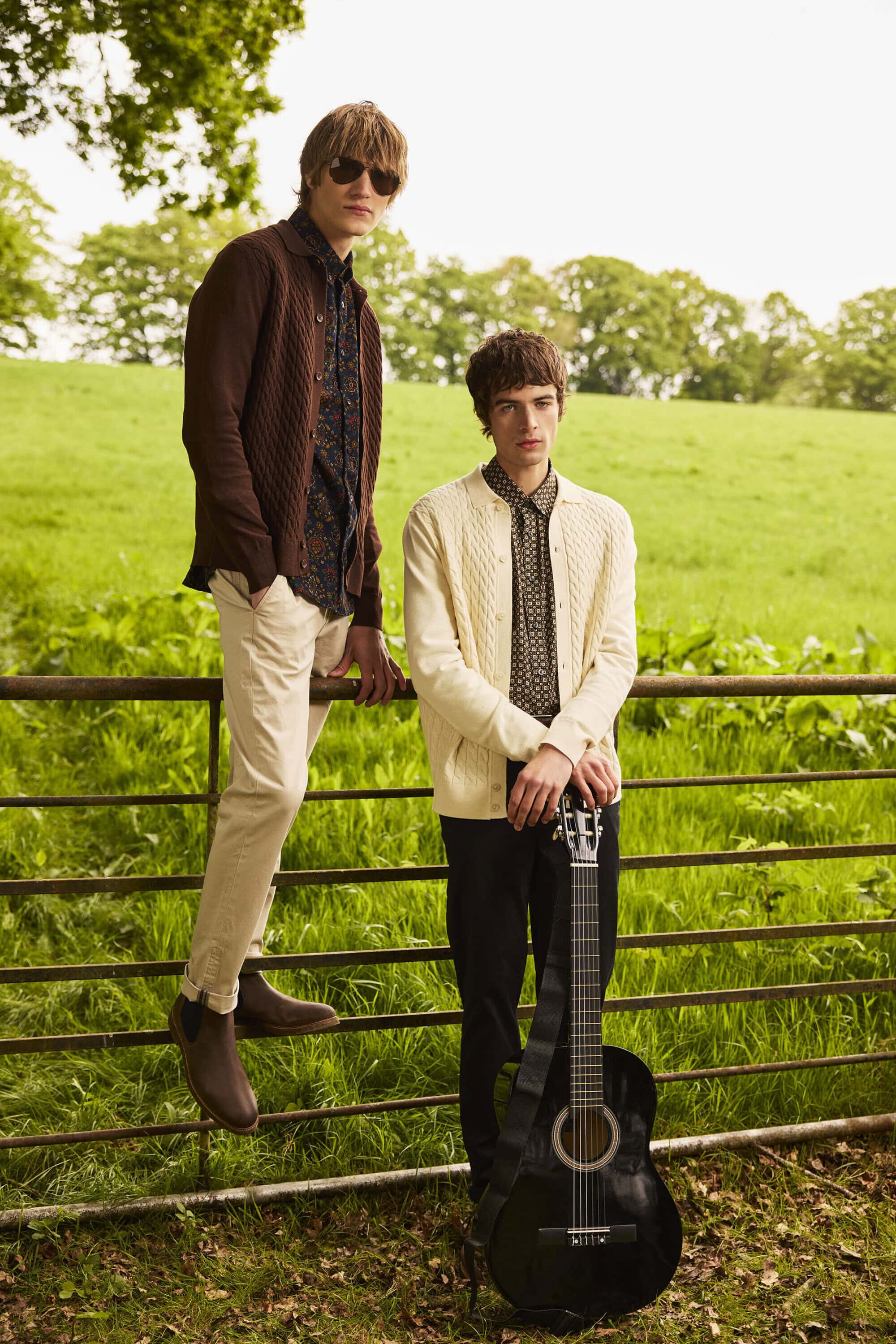Two young male models in knit cardigans and patterned shirts posing by a metal gate in a green meadow.