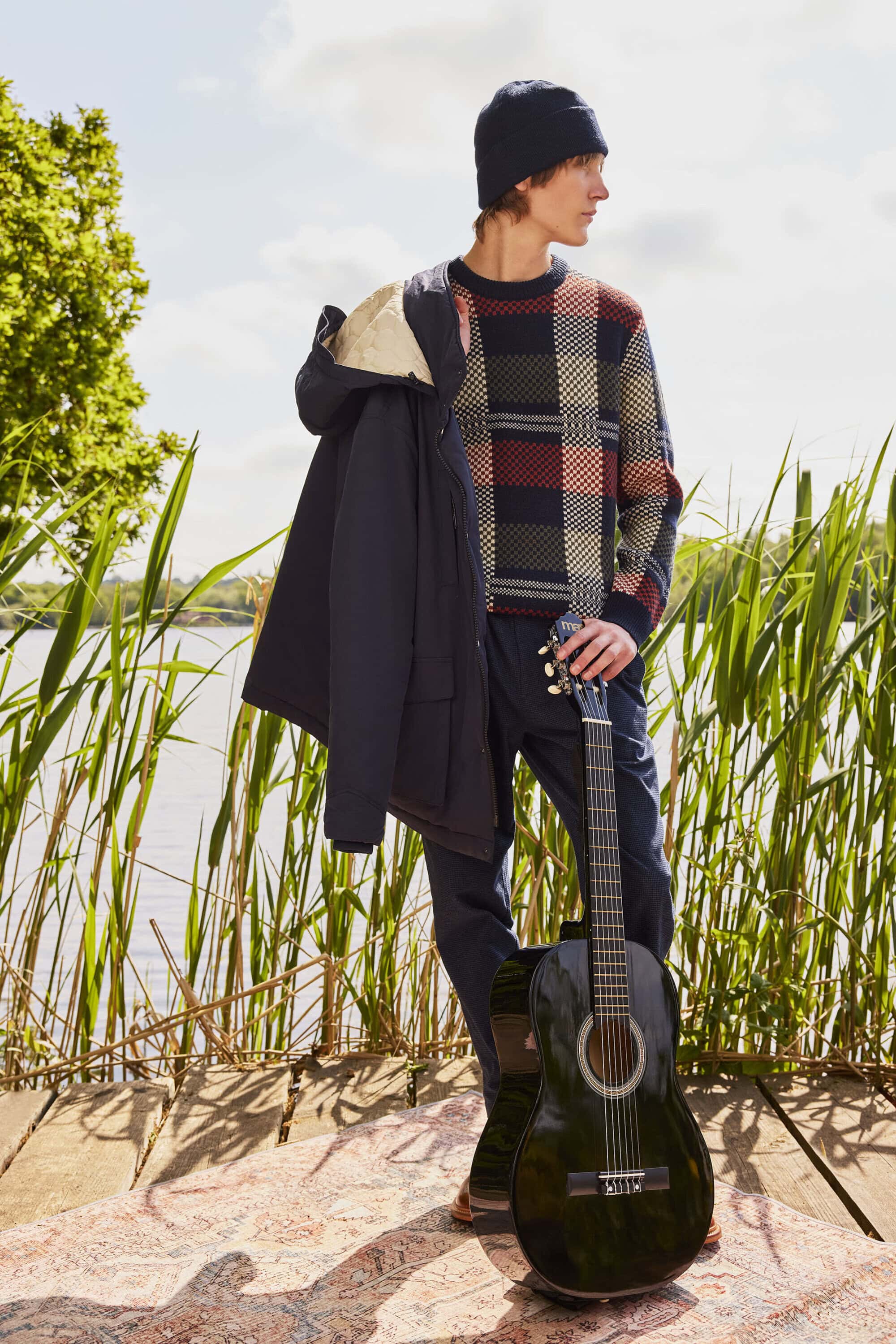 Model wearing a patterned knit sweater and beanie holding an acoustic guitar by a lakeside.