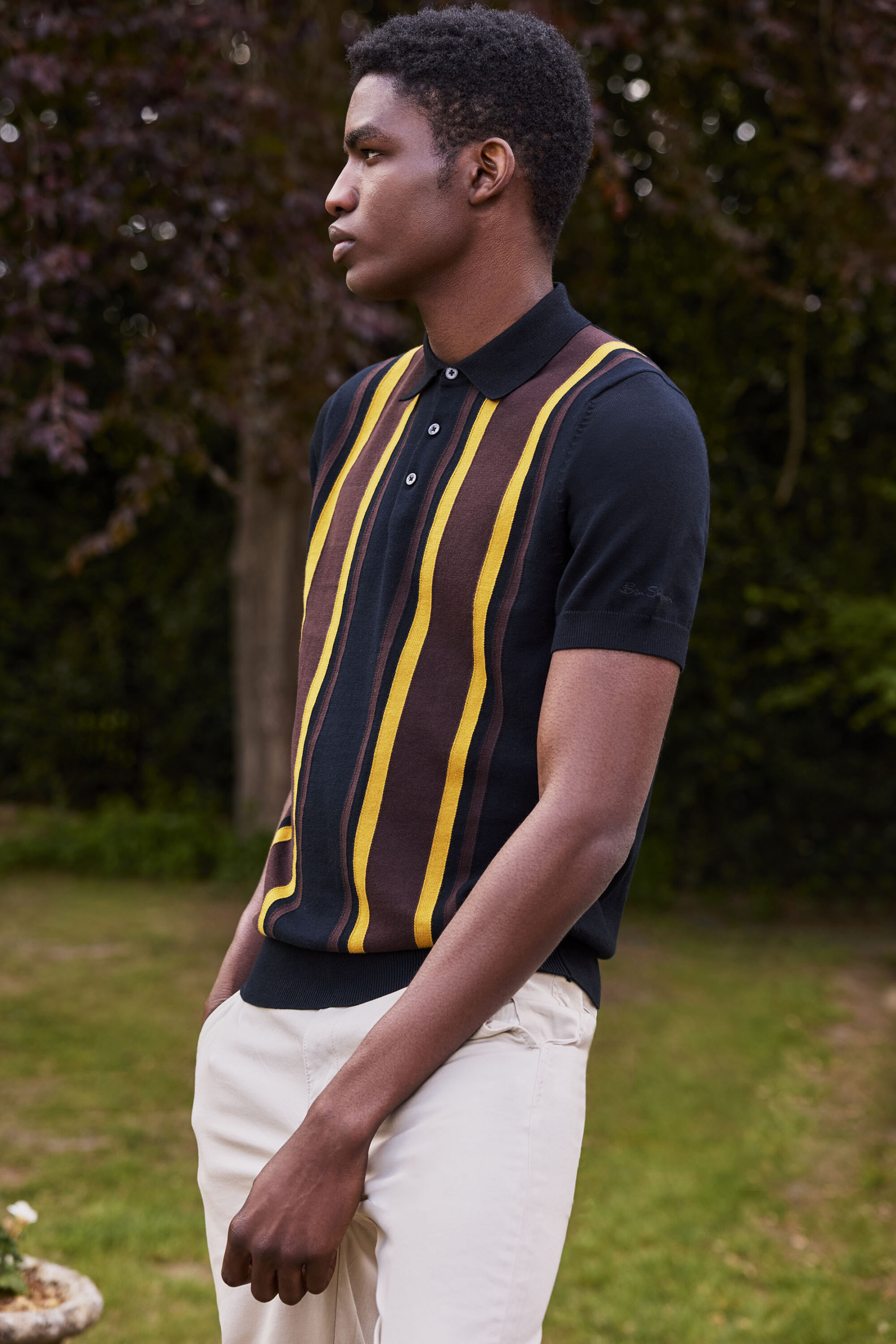 Black man wearing a striped knit polo shirt and light trousers outdoors in a park setting