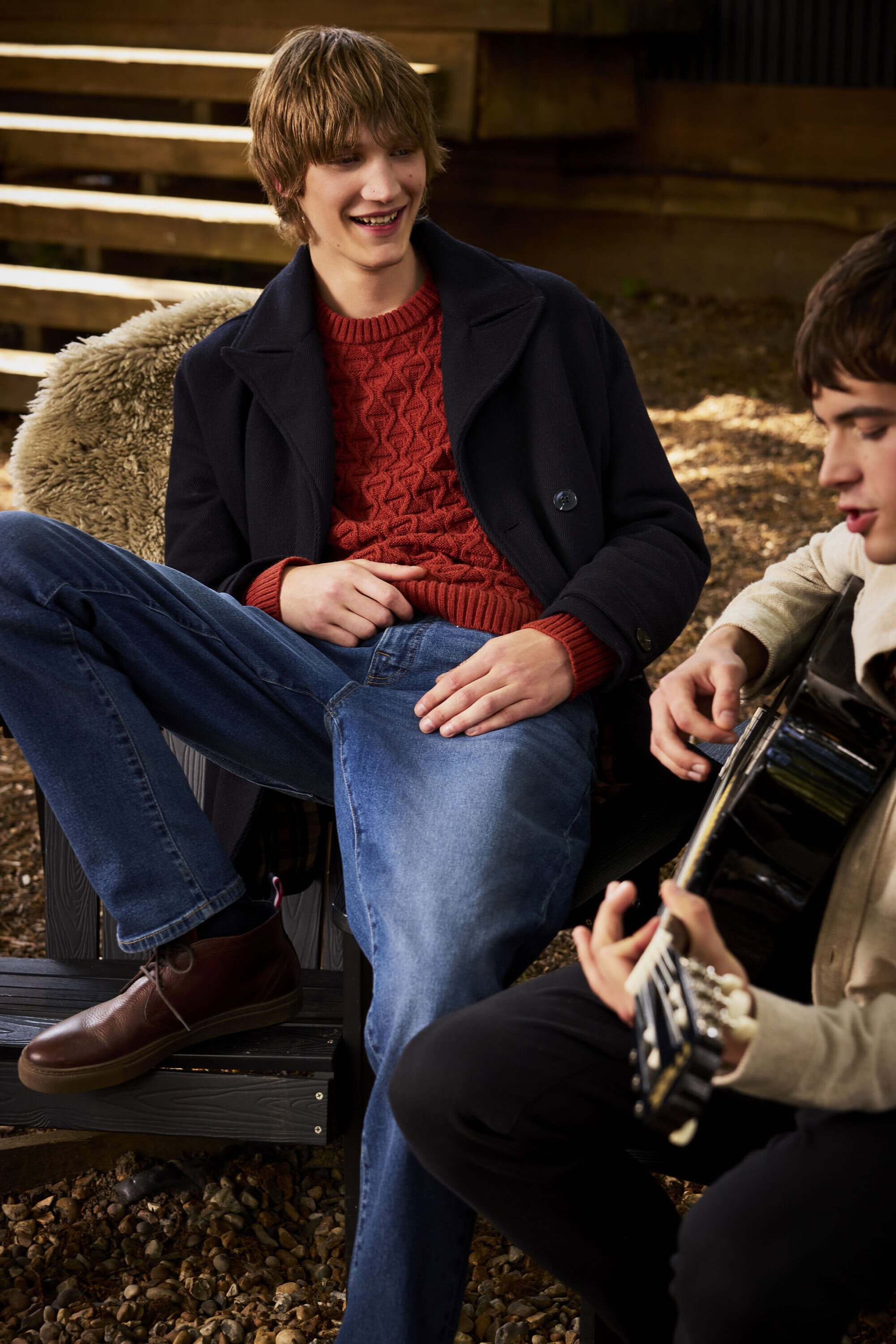 Two young men seated outdoors, one smiling in a red cable-knit sweater and dark jacket playing guitar.