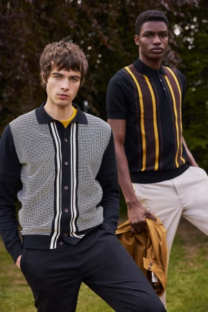 Two male models wearing patterned knit shirts and polo shirts posing outdoors in a park by trees.
