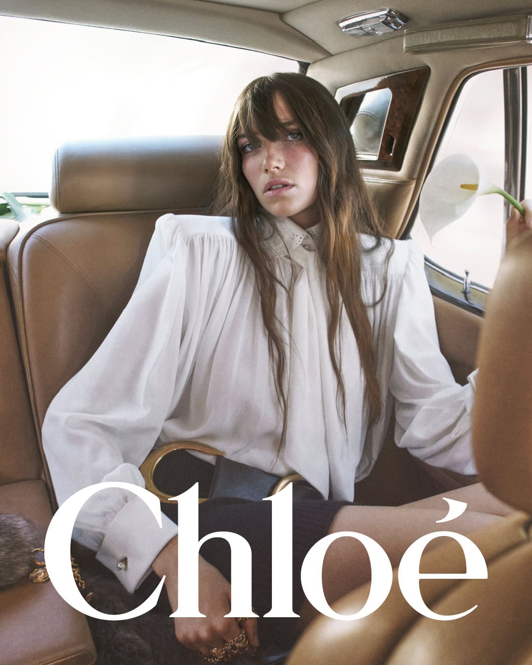 Model with long brown hair wearing a white blouse and dark skirt seated inside a luxury vintage car interior