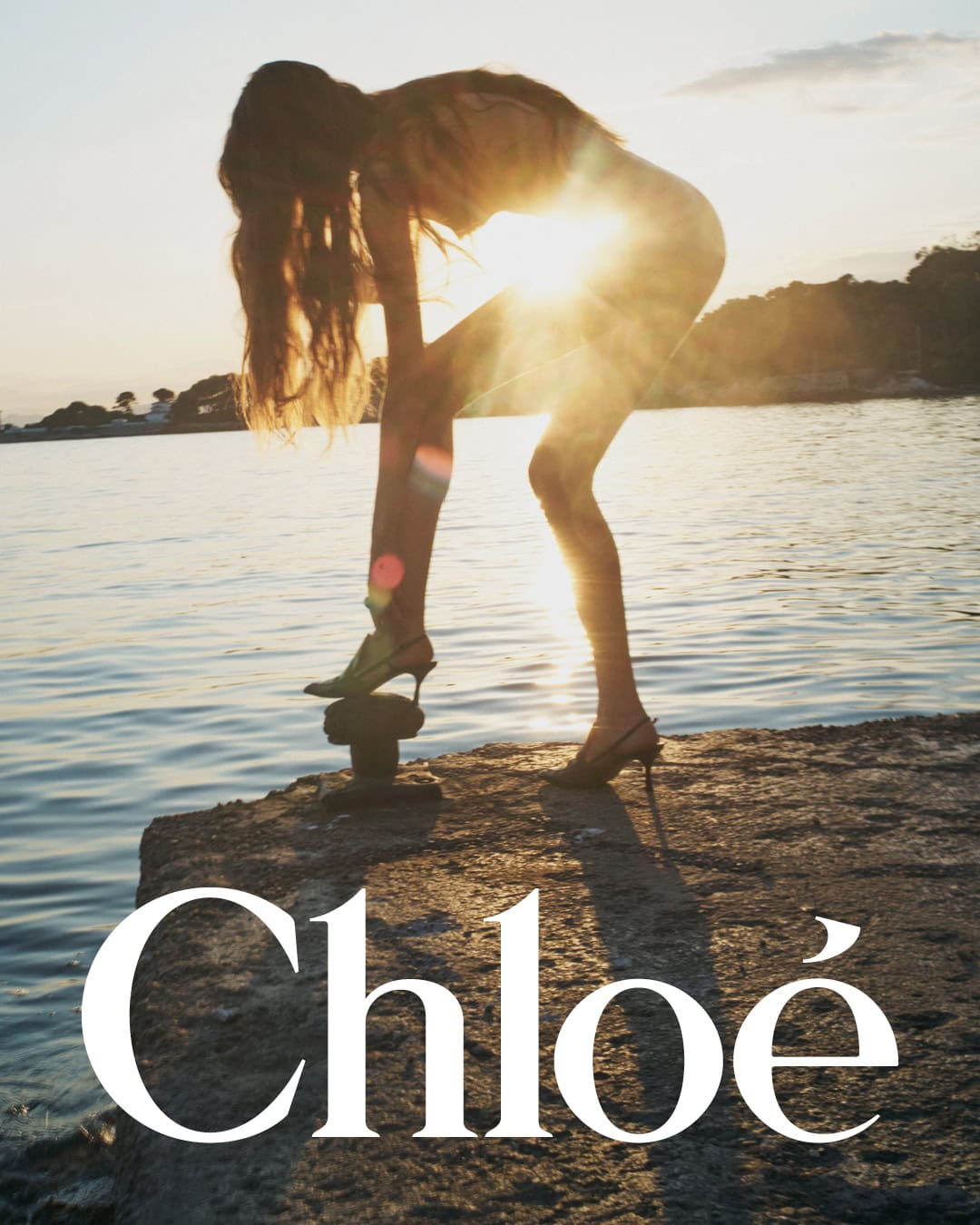 Model standing on a stone pier by the water at sunset with the Chloé logo overlaid at the bottom