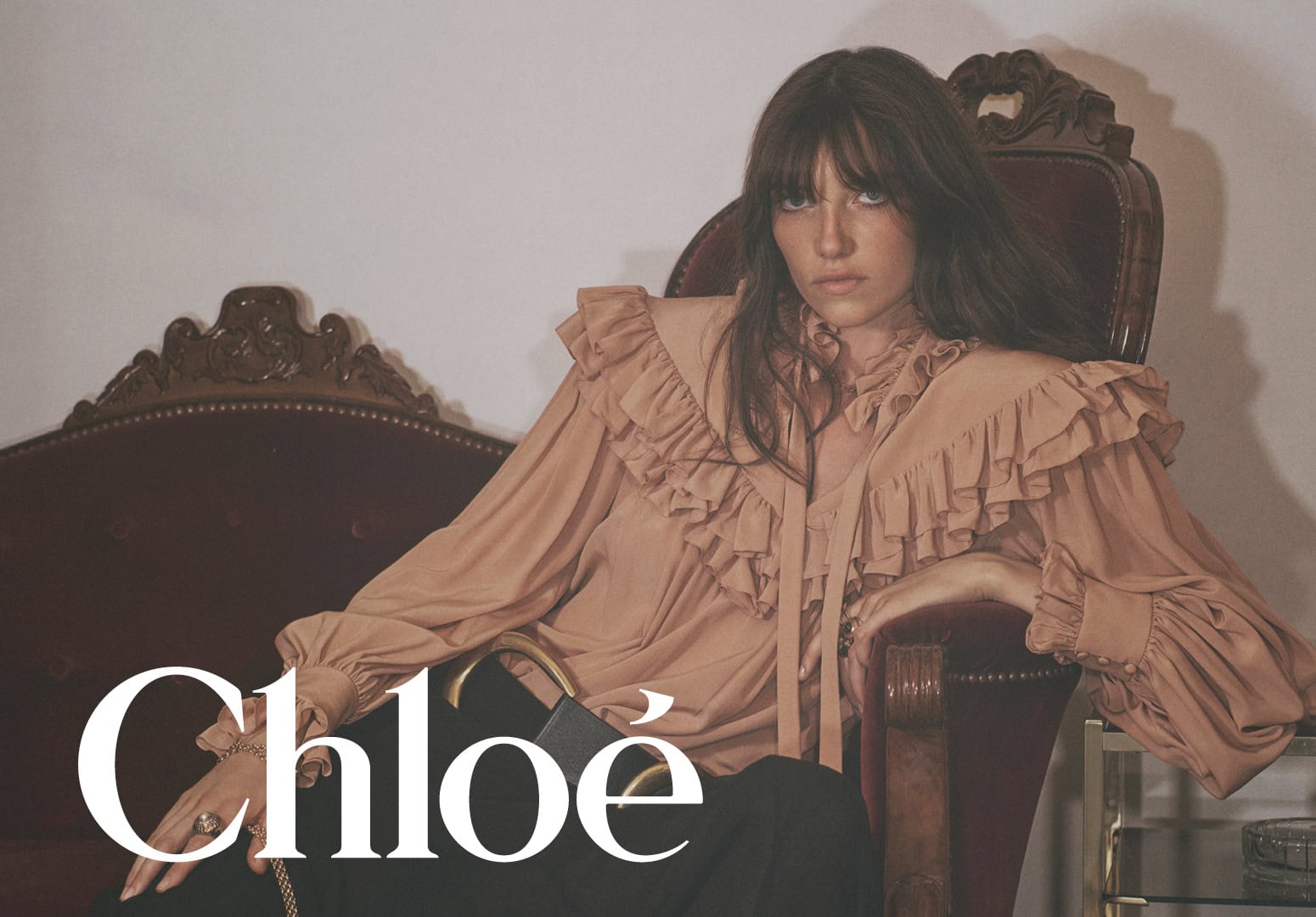 Model Grace Hartzel seated on a velvet chair wearing a ruffled blouse for the Chloe Winter 2025 ad campaign.