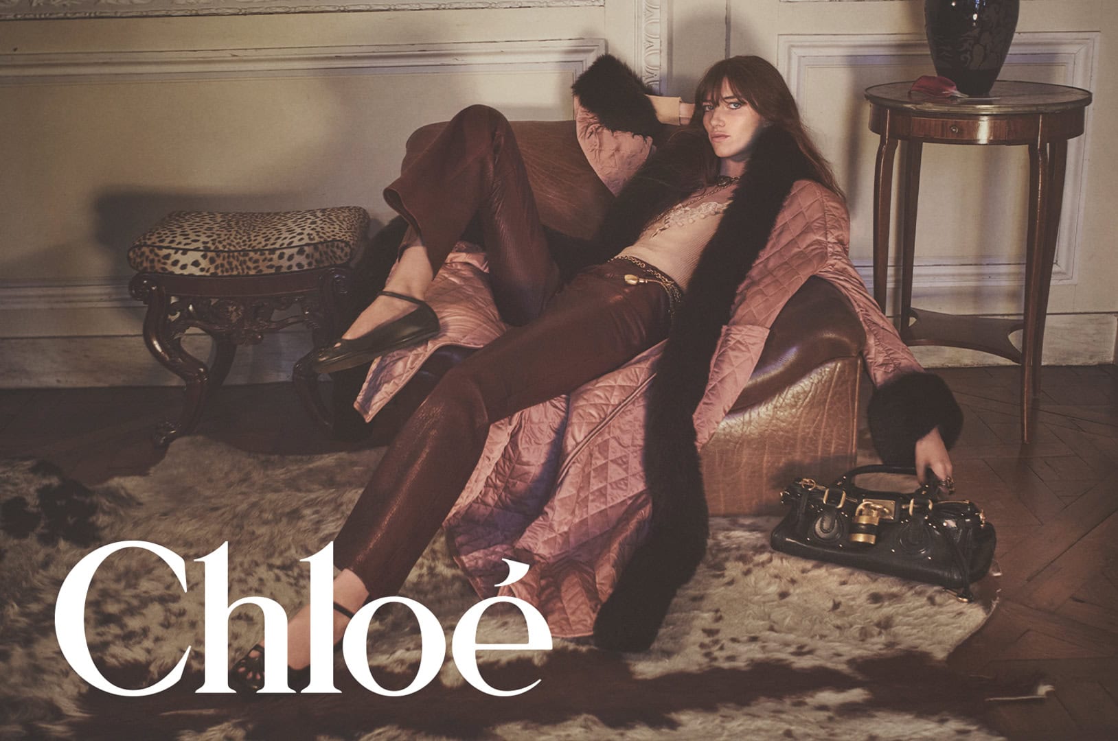 Model Grace Hartzel reclining in an armchair wearing a quilted coat and leather pants with a dark handbag for Chloé.