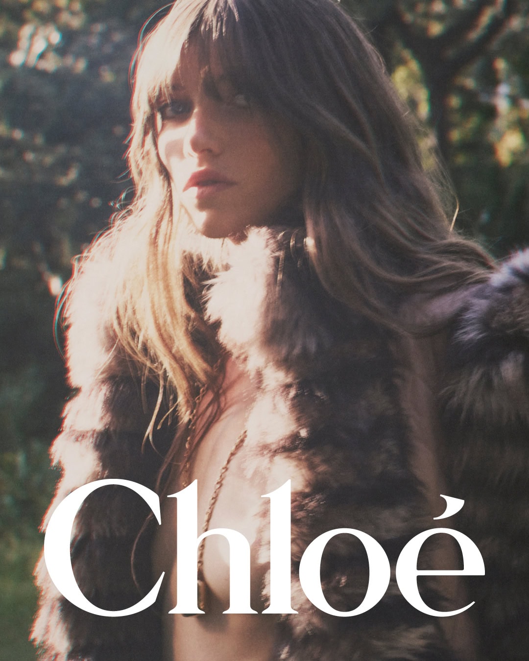 Model with fringe hair wearing a textured fur vest outdoors against a soft green tree foliage background.