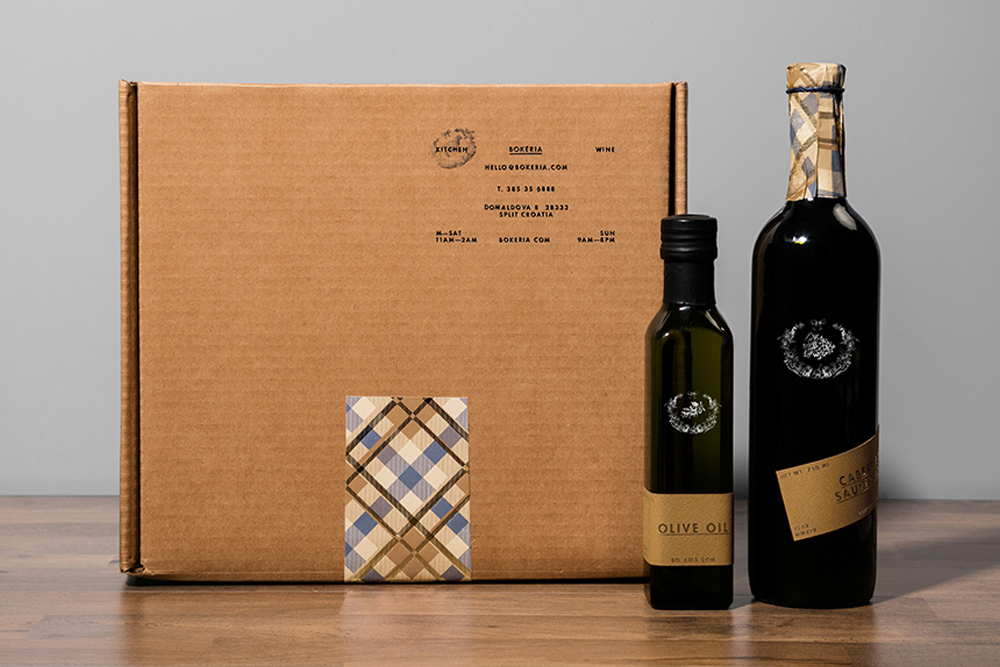 Brown cardboard shipping box with brand typography beside a tall wine bottle and a smaller olive oil bottle on a wooden table.