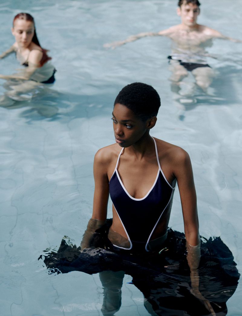 Three people in a swimming pool, one African woman in the foreground wearing a black one-piece swimsuit with white trim, waist deep in water.