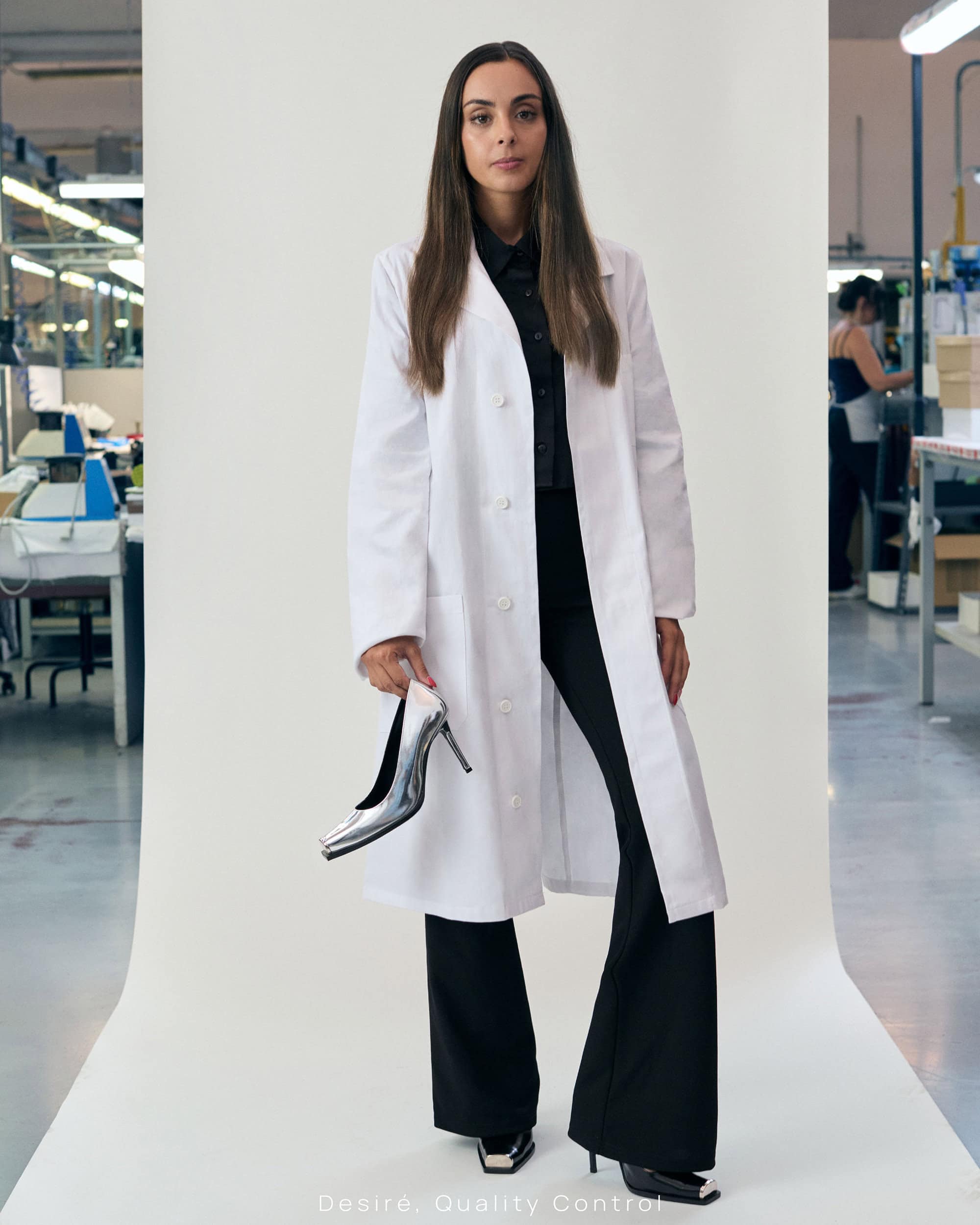 Woman in white lab coat holding a metallic high-heel shoe in front of a white seamless backdrop with an industrial workshop visible