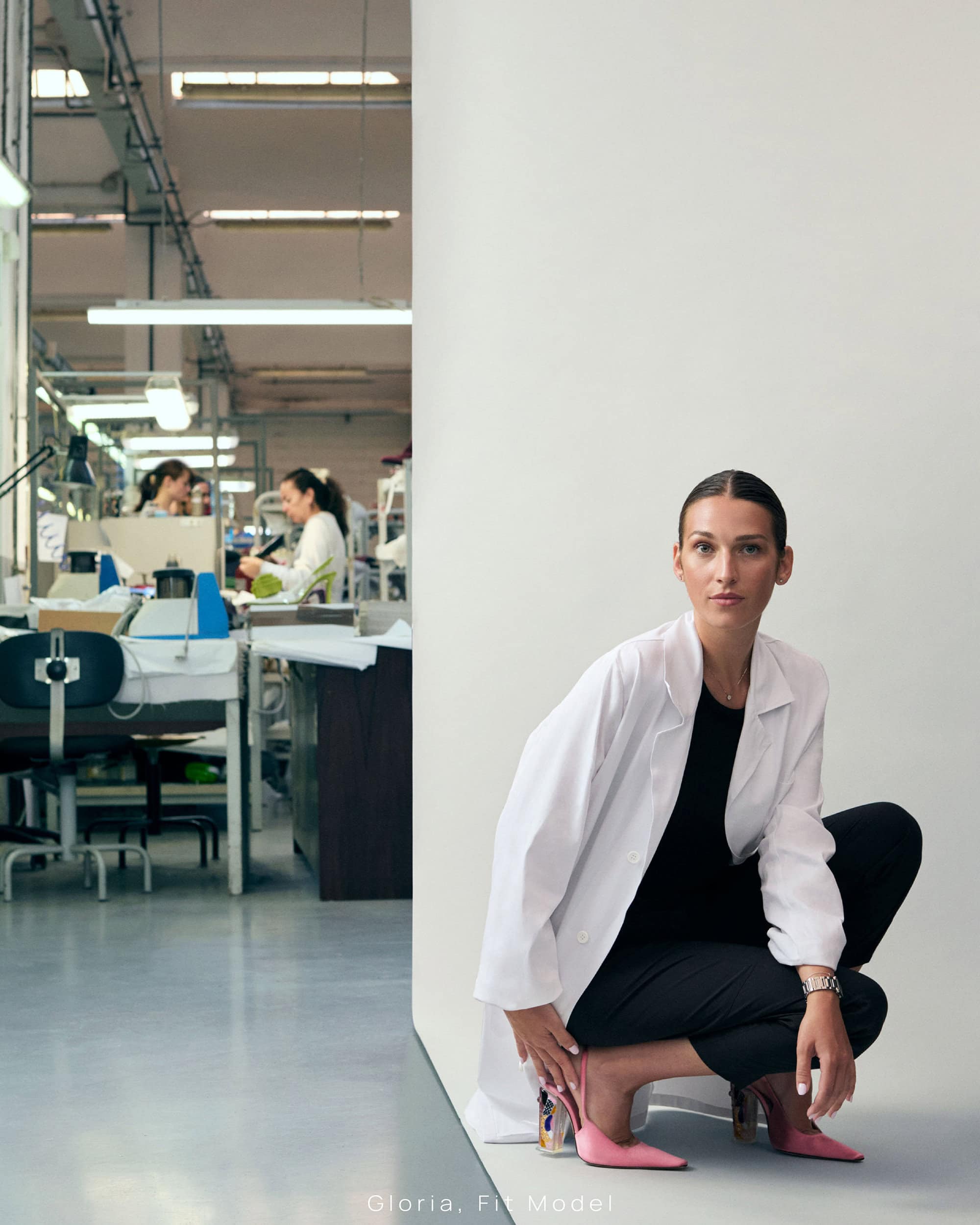 Woman in white coat and black trousers crouching on a white floor in a factory setting