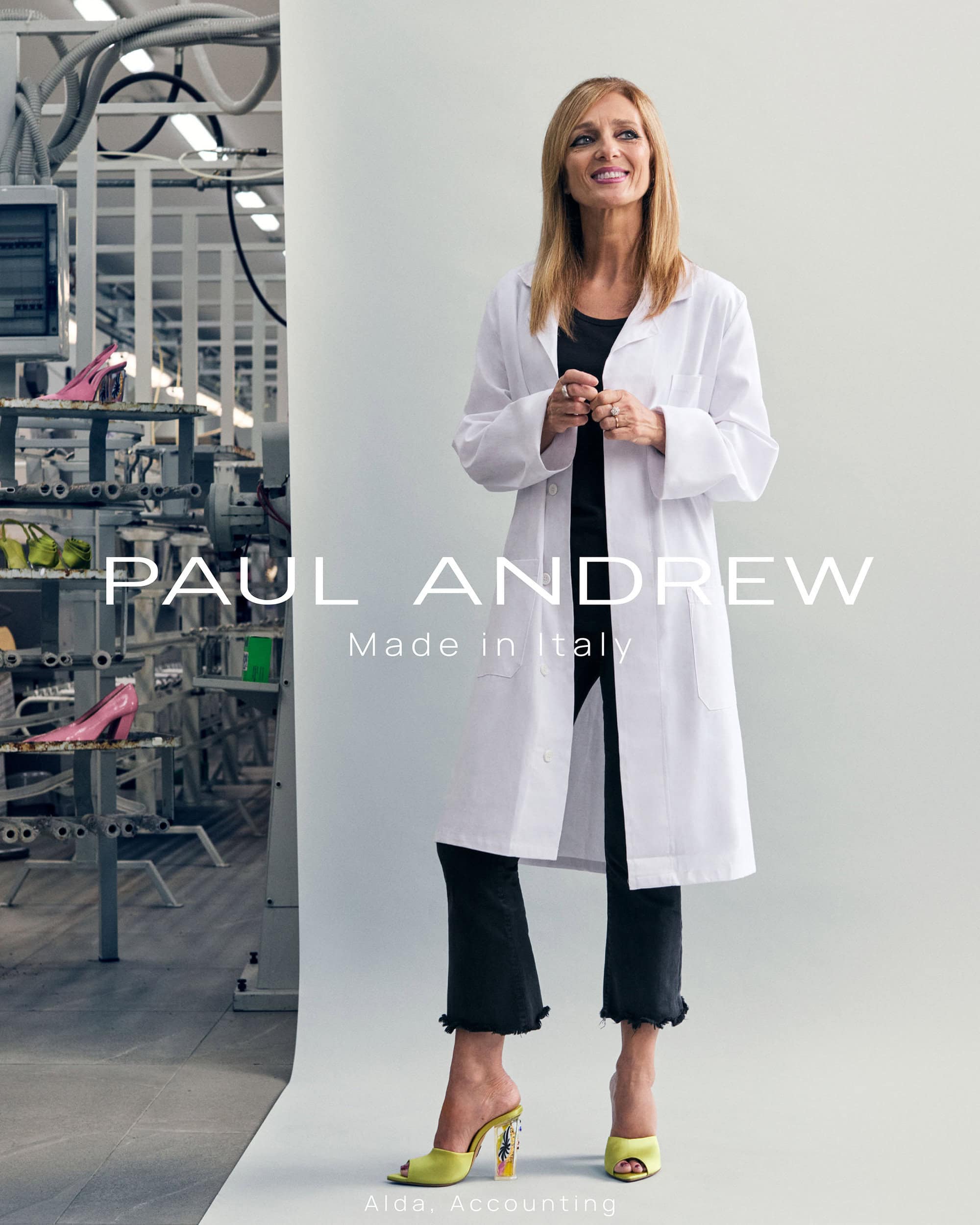 Woman in a white coat standing in front of a white seamless background with industrial shelving visible on the left.