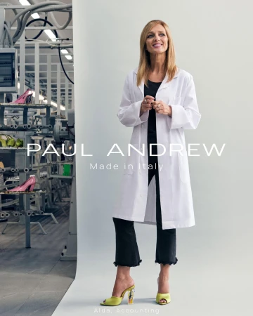 Woman in a white coat standing in front of a white seamless background with industrial shelving visible on the left.