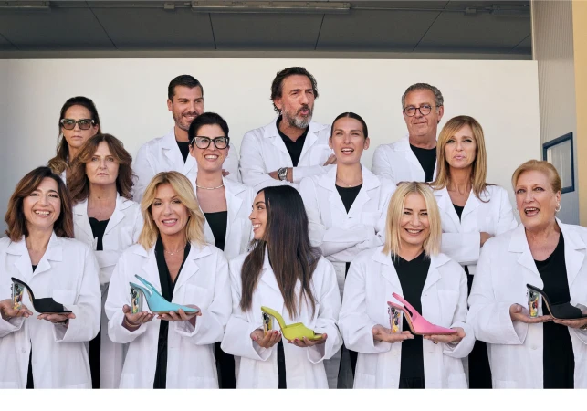 Group of twelve people wearing white lab coats holding high-heeled shoes against a plain white studio backdrop.