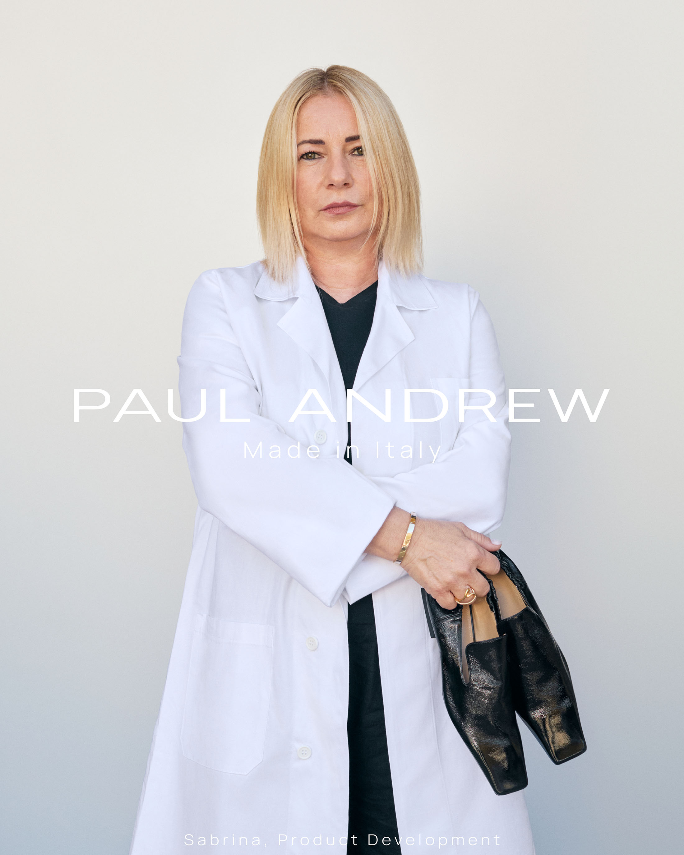 Blonde woman wearing a white coat holding black leather loafers against a plain background for Paul Andrew Season Seven ad campaign