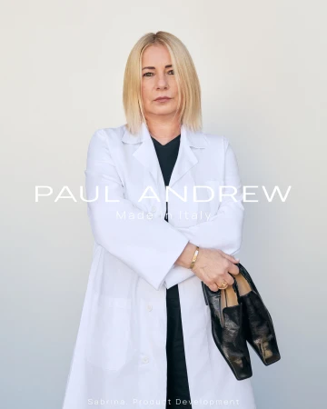 Blonde woman wearing a white coat holding black leather loafers against a plain background for Paul Andrew Season Seven ad campaign