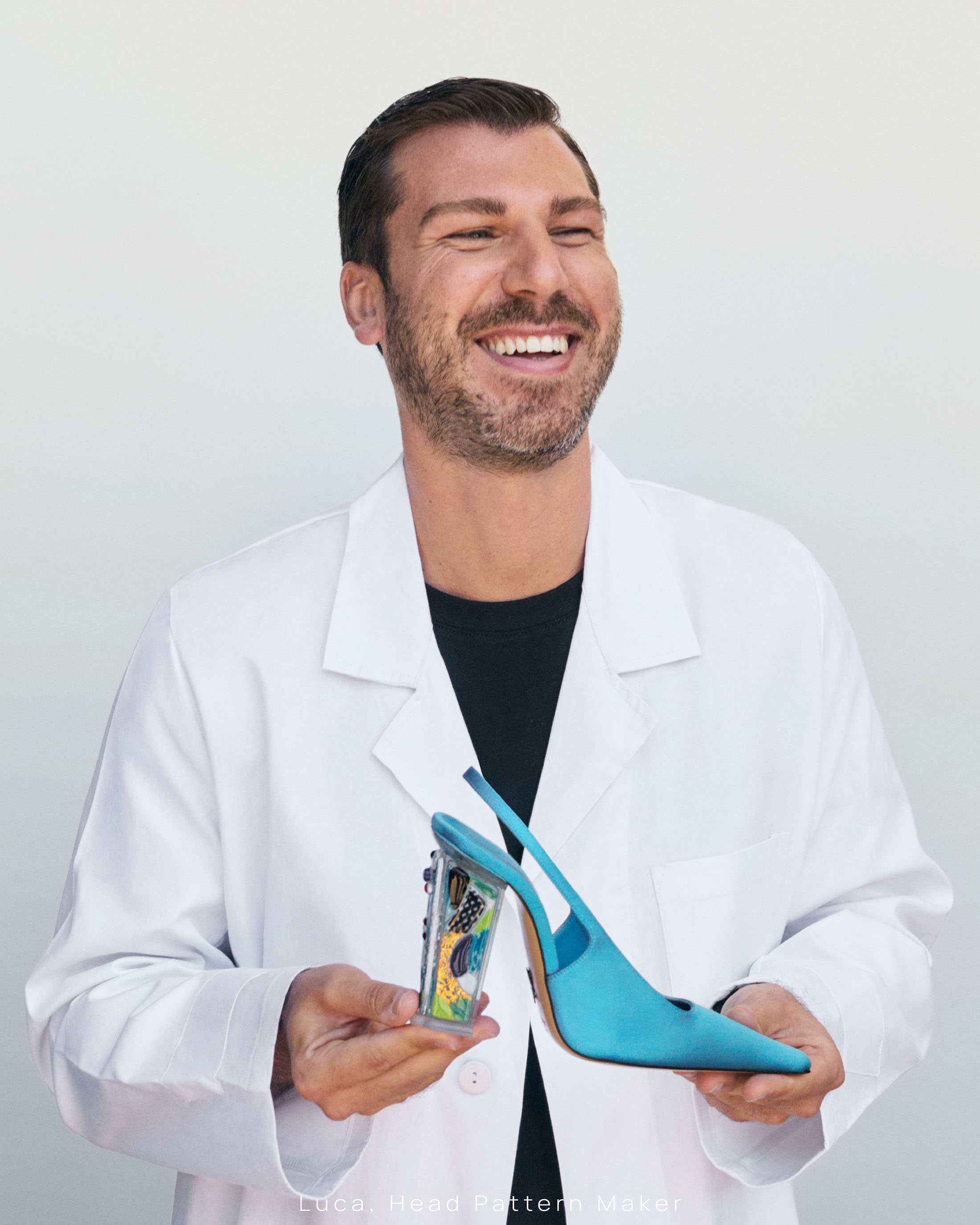 Smiling man wearing white coat holding a bright blue slingback shoe against a pale background