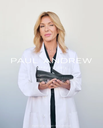 Blonde woman in a white lab coat holding a black leather loafer against a light grey studio background
