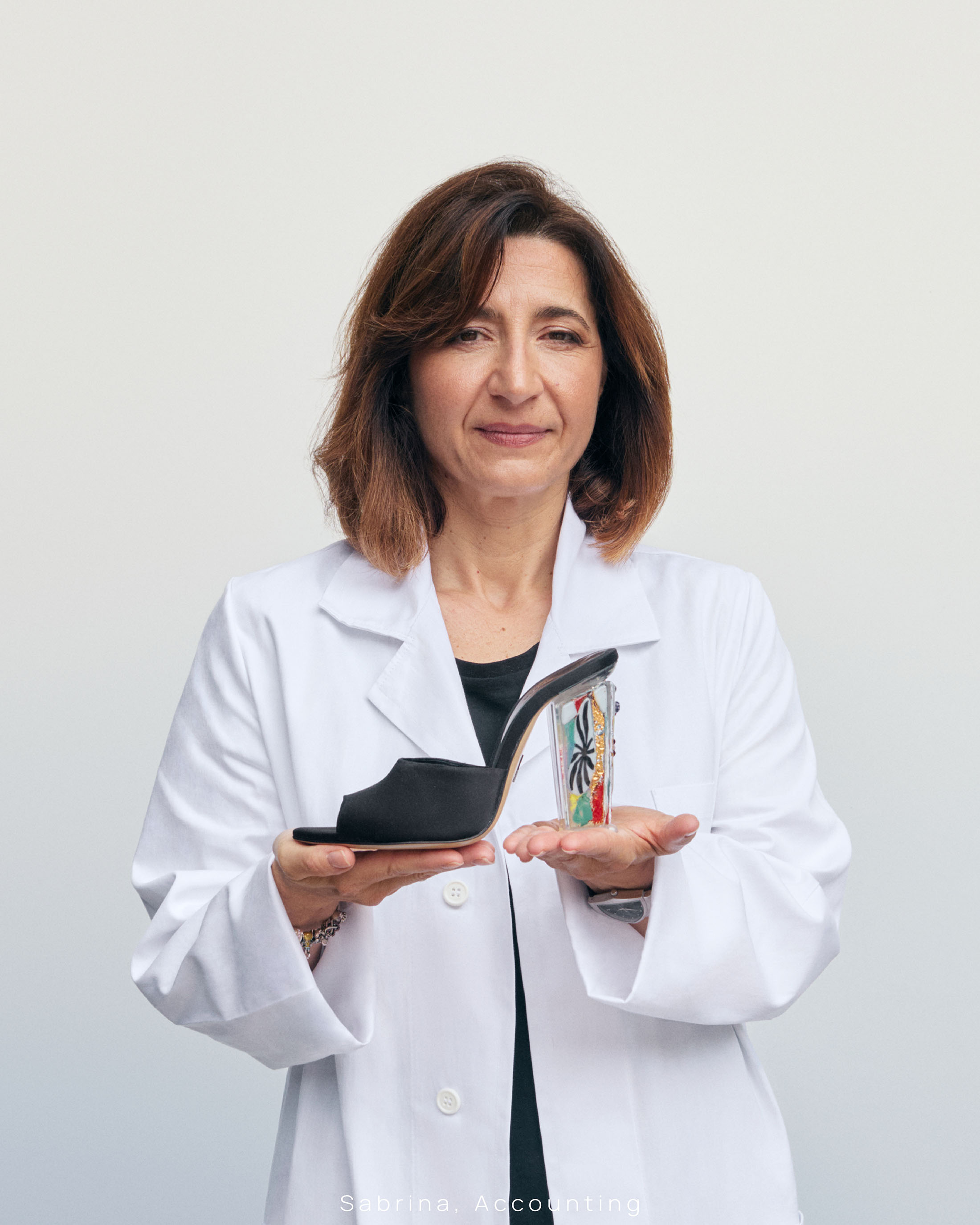 Middle-aged woman in a white lab coat holding a black high-heeled shoe with a patterned clear heel against a light grey background