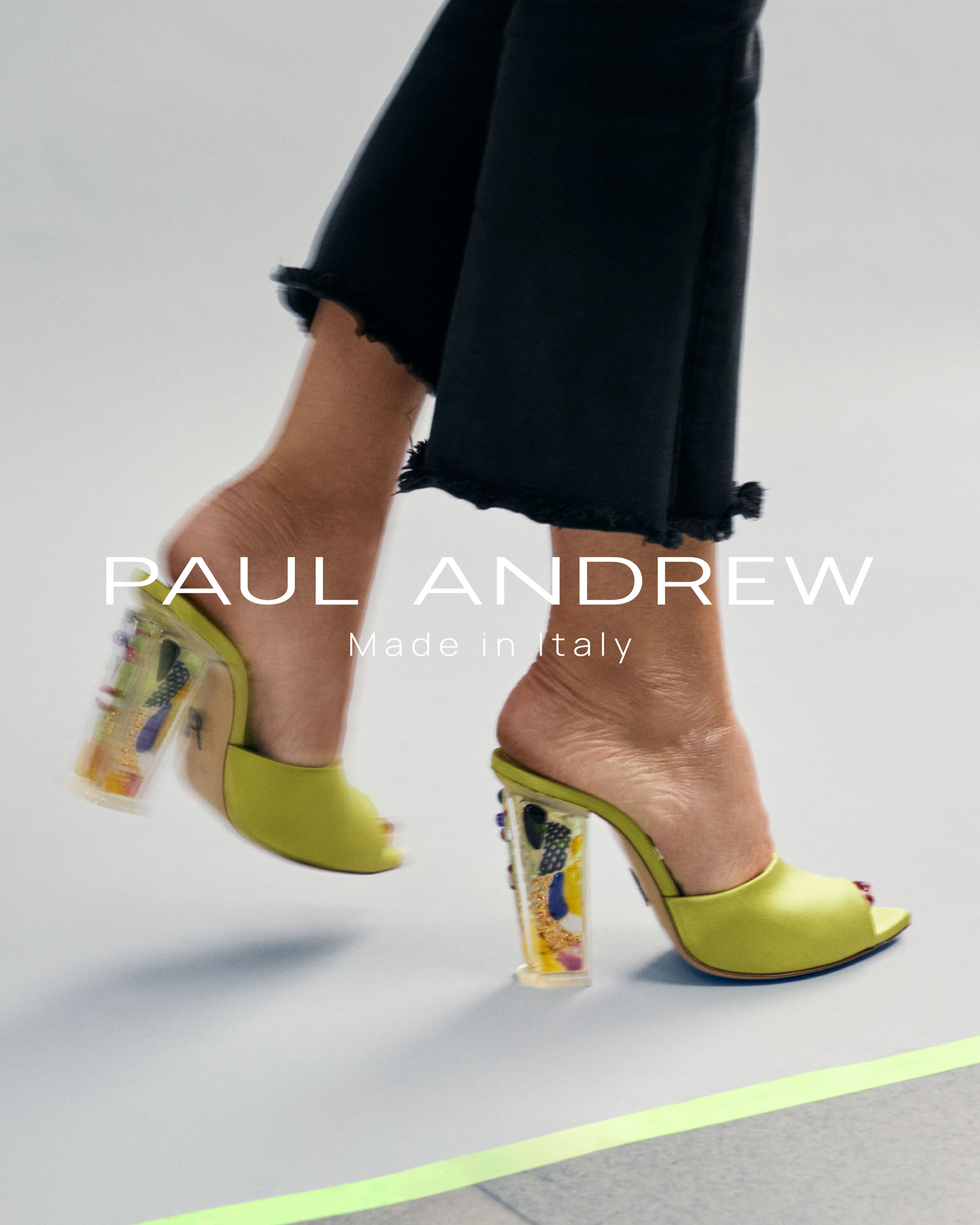 Close-up of feet wearing chartreuse heeled mules with patterned transparent heels and black frayed hem trousers against a light grey background.