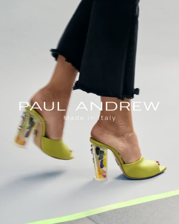 Close-up of feet wearing chartreuse heeled mules with patterned transparent heels and black frayed hem trousers against a light grey background.