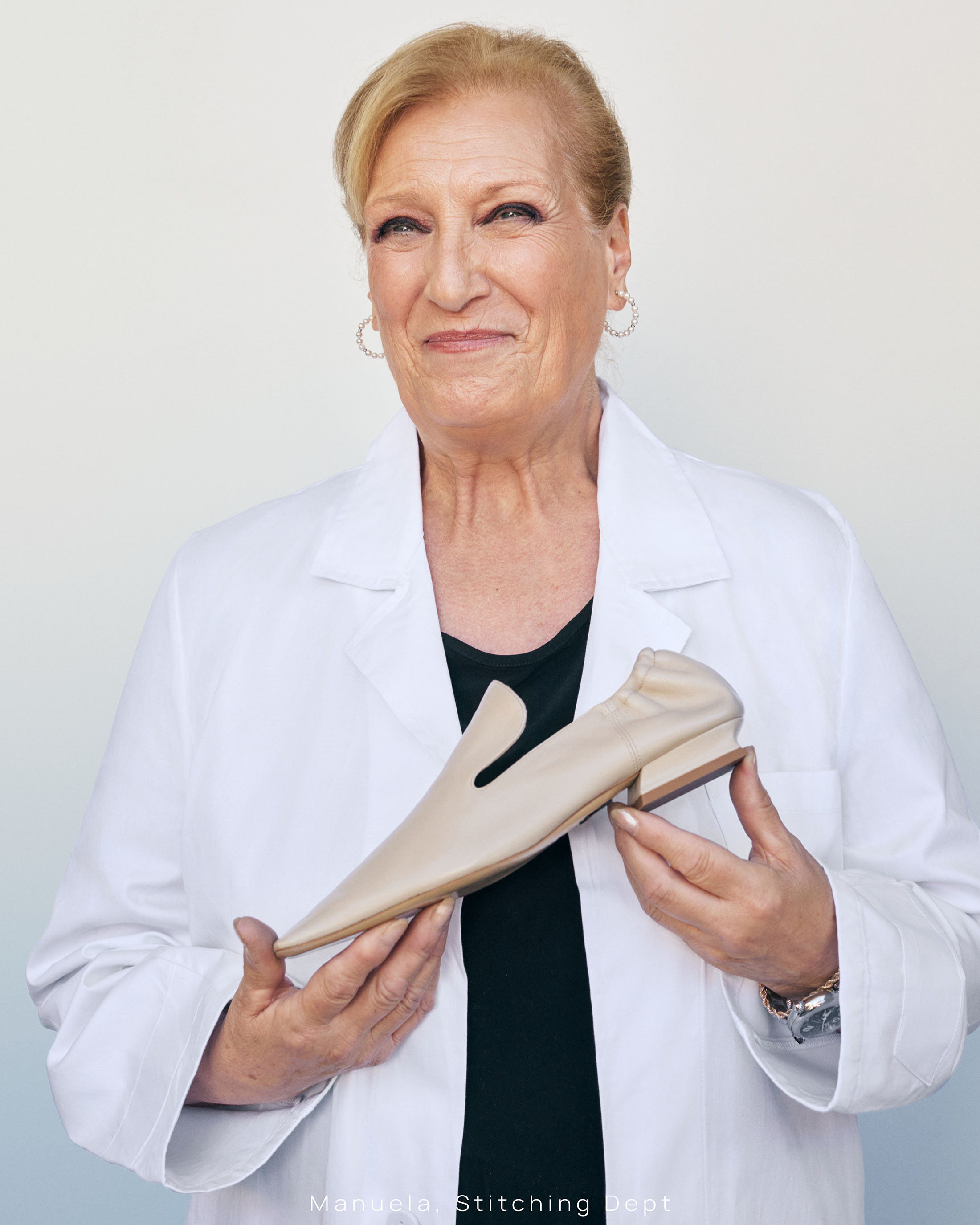 Elderly woman wearing a white coat holding a leather shoe against a neutral background for Paul Andrew Season Seven campaign