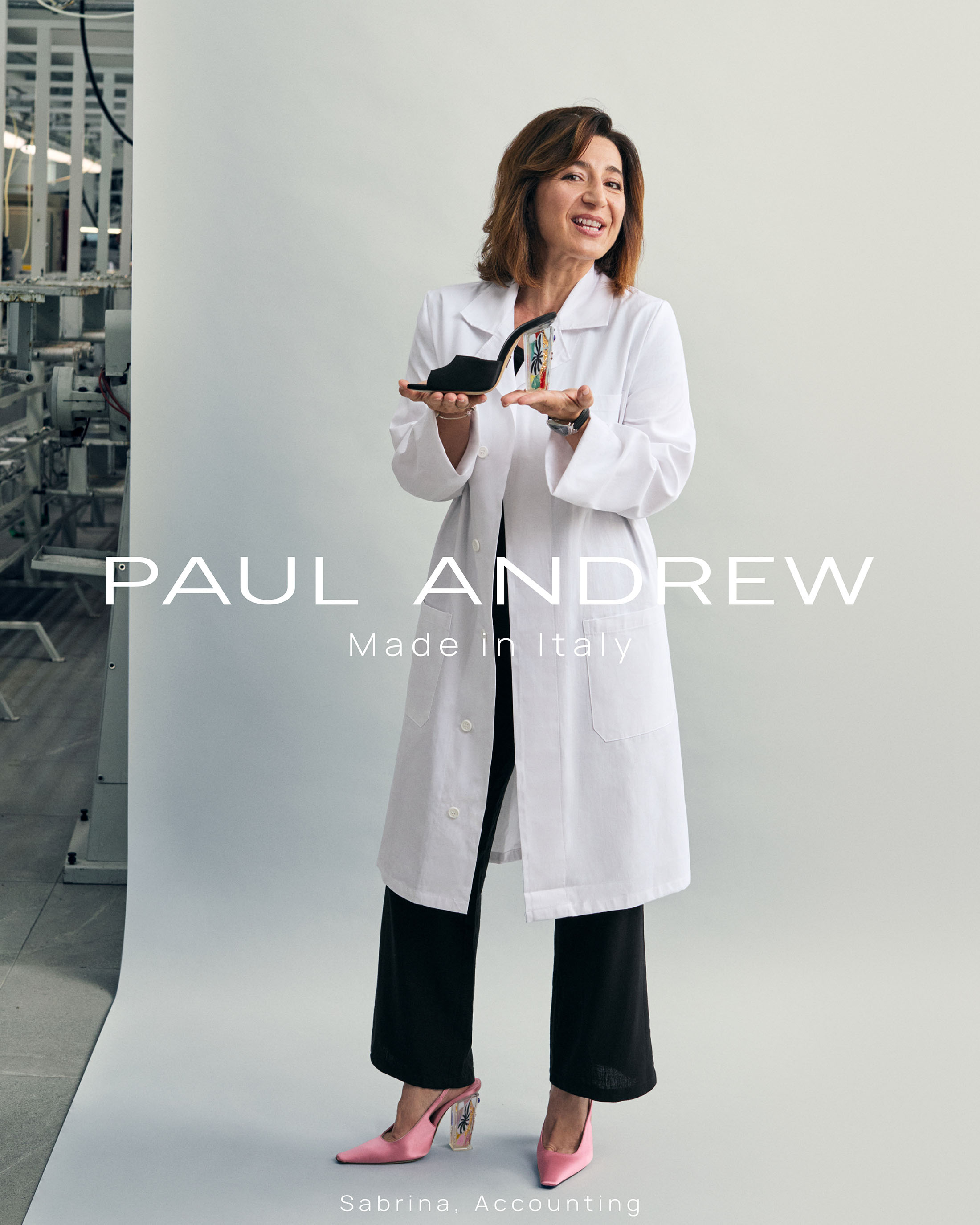 Woman wearing white lab coat over black trousers holding a black high heel shoe in a studio setting