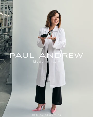 Woman wearing white lab coat over black trousers holding a black high heel shoe in a studio setting