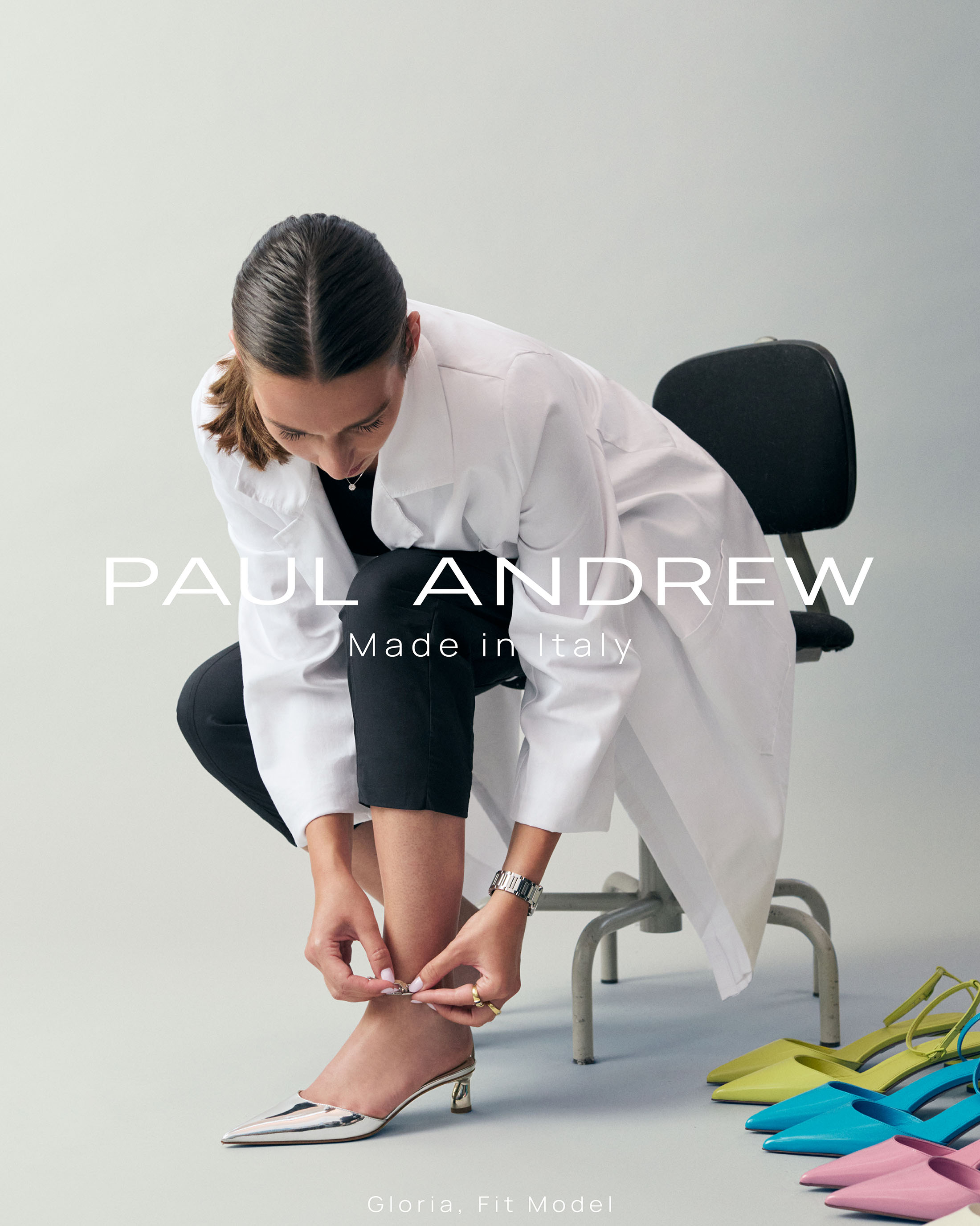 Model wearing white coat fastening silver pointed heel shoe in studio for Paul Andrew ad campaign