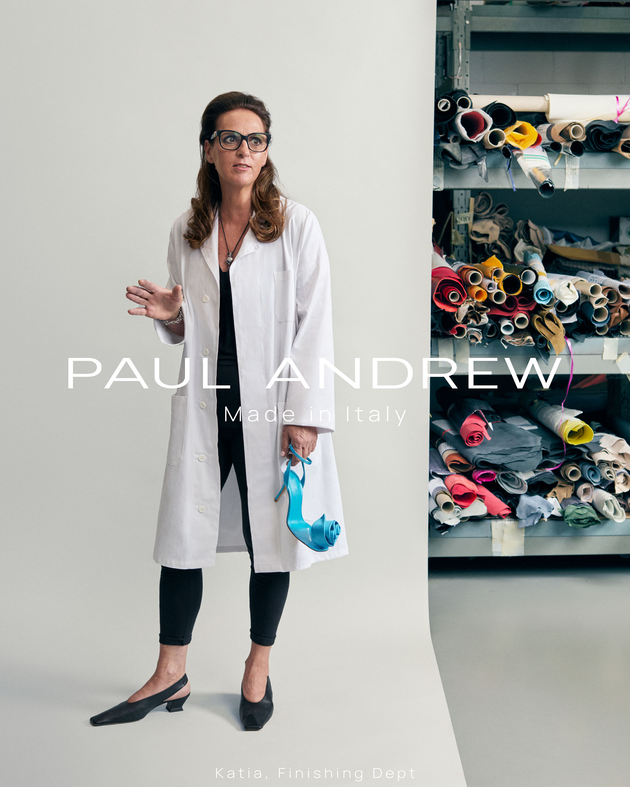 Woman wearing glasses and a white lab coat holding a blue shoe in a workshop setting for Paul Andrew