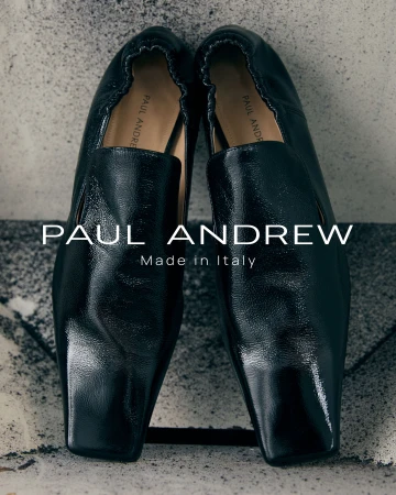 Close-up product shot of a pair of glossy black leather loafers placed upright against a textured stone background.